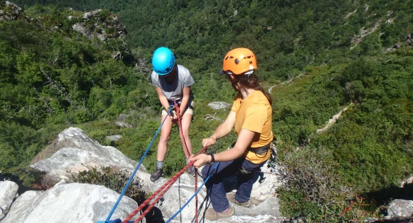 Two people wearing safety gear are secured by ropes near the edge of a cliff. One person appears to be an instructor, giving direction to the other person. Below them is a green wooded area. 